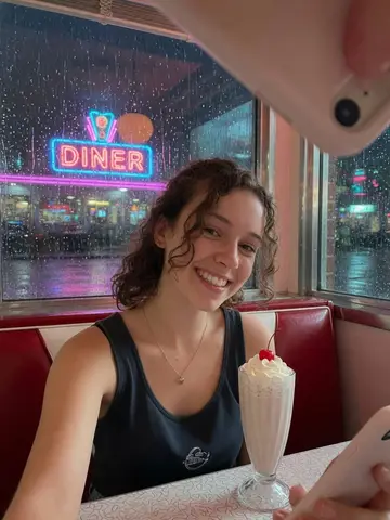 Exempel på Neondiner: Personen sitter i ett bås på en retrodiner från 1950-talet en kväll, med neonsken, en milkshake på bordet och regn på fönstret.